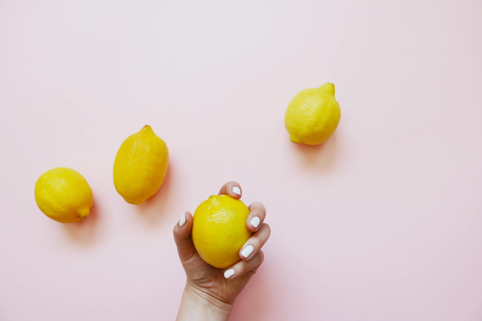 A hand holding a fresh lemon on a soft pink background with additional lemons nearby, symbolizing renewal and fresh starts.