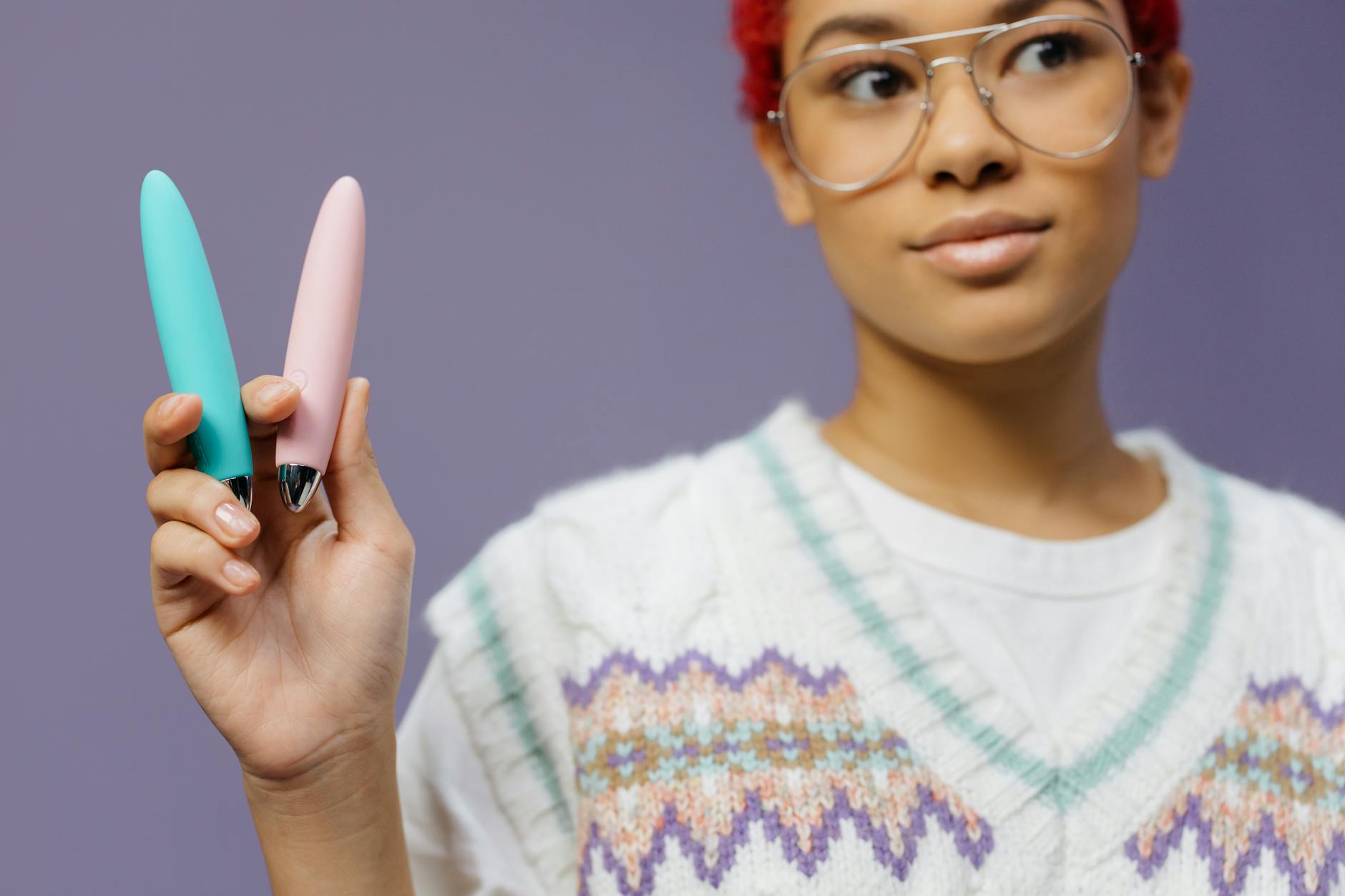 A woman in eyeglasses holding colorful lemon vibrators and clitoral massagers against a purple background.